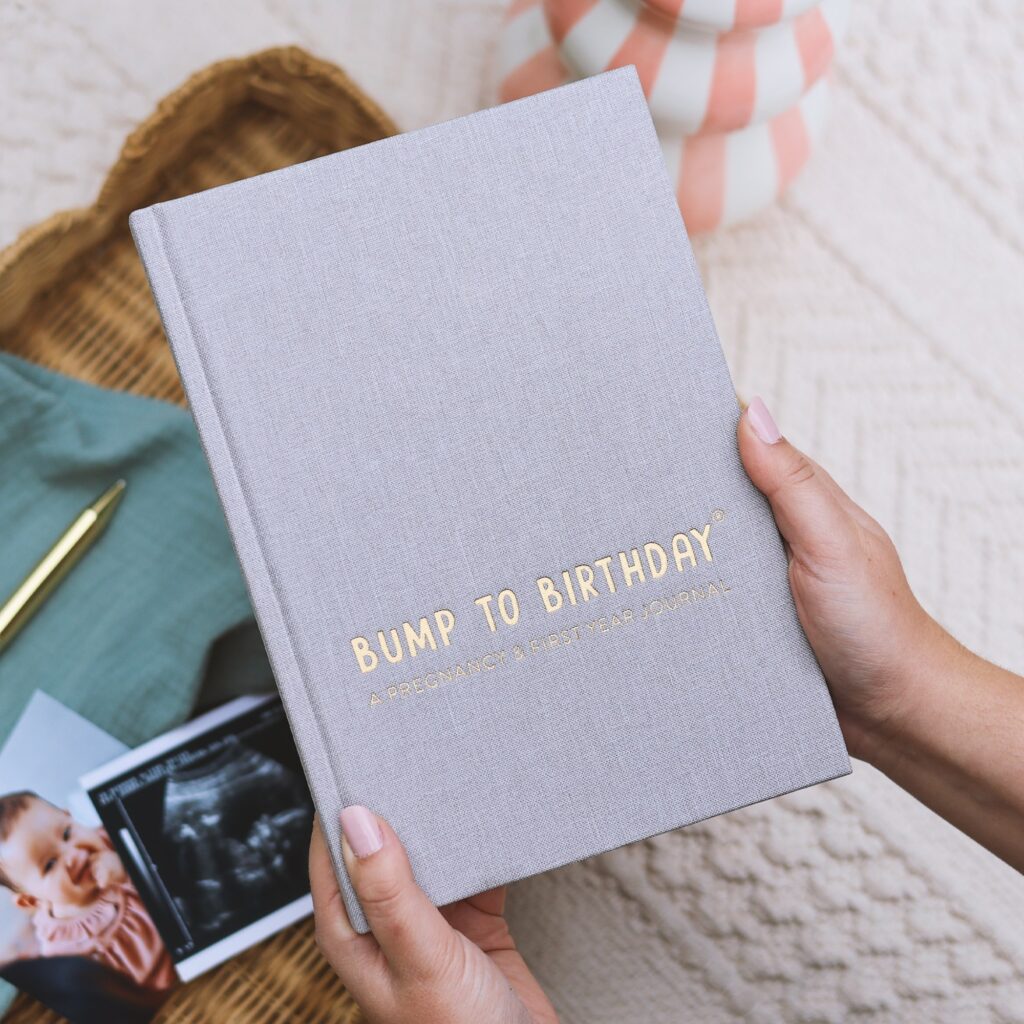 Holding grey journal, with gold foil lettering saying "Bump To Birthday". There is a baby scan and baby photo in the background.