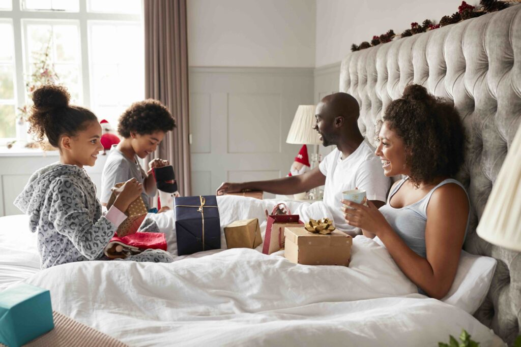 Young kids opening gifts on parents bed on Christmas morning while their parents sit up in bed watching, side view.