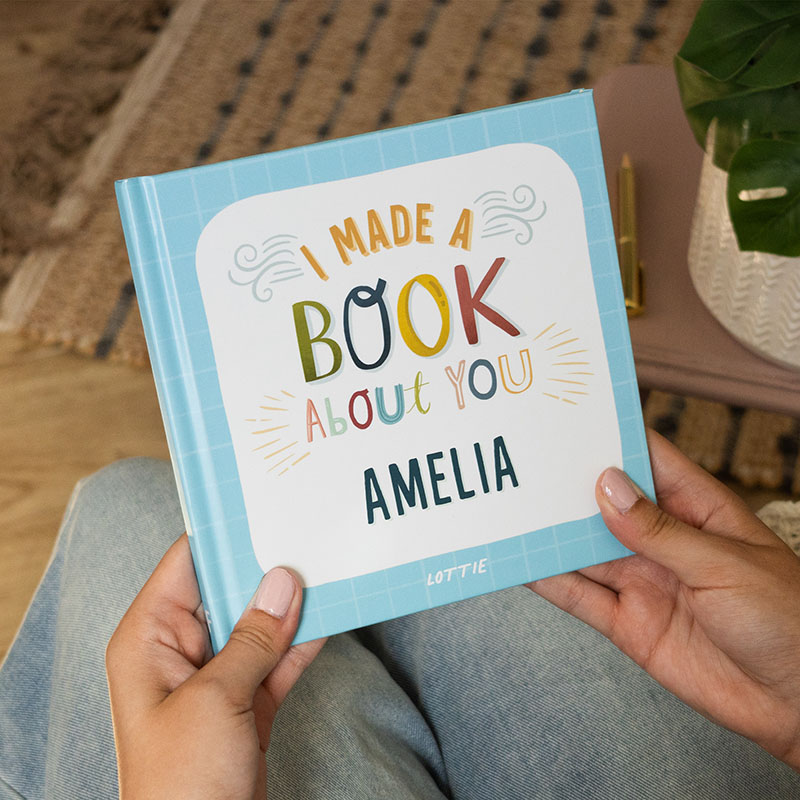 Woman's hands holding a light blue "I Made a Book About You Amelia" friendship book. She is sat in a cosy environment ready to read the book.