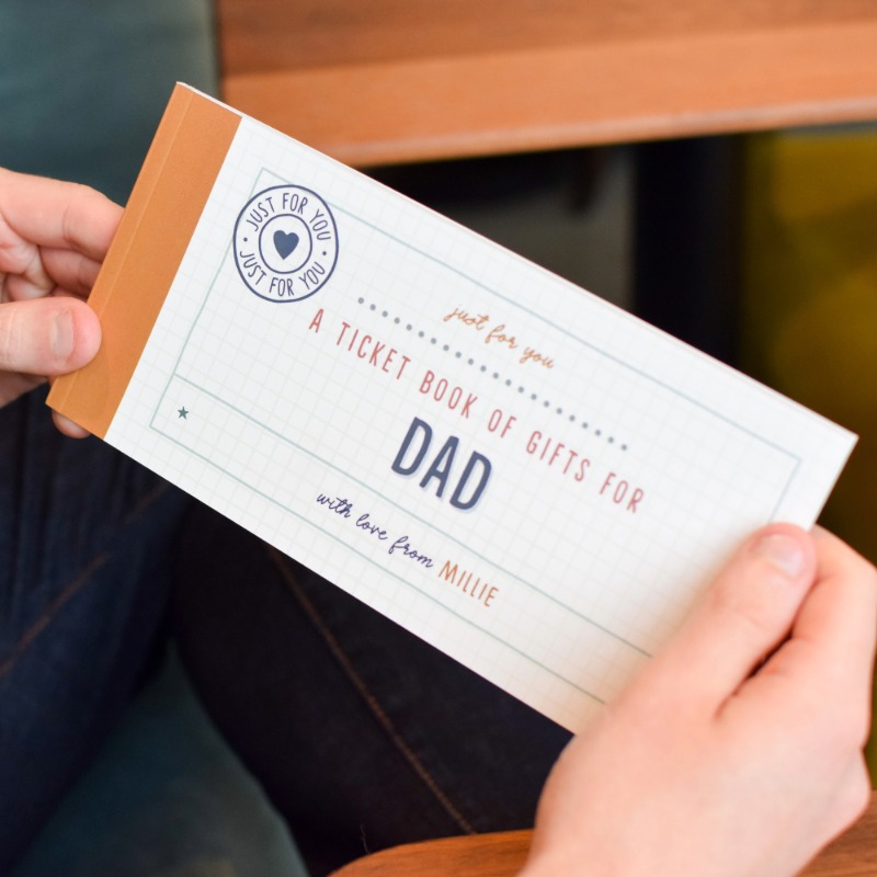 Man holding large ticket book with a brown spine. "A ticket book of gifts for Dad. With love from Millie". The book features a playful, yet sophisticated design. The gift is being received in a warm masculine environment.