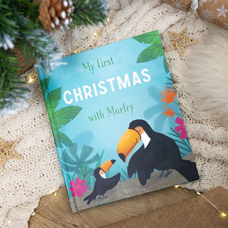 My First Christmas With Marley baby book on a cream throw, surrounded by Christmas ornaments and a tree. The book features a baby and adult toucan reaching for each other.