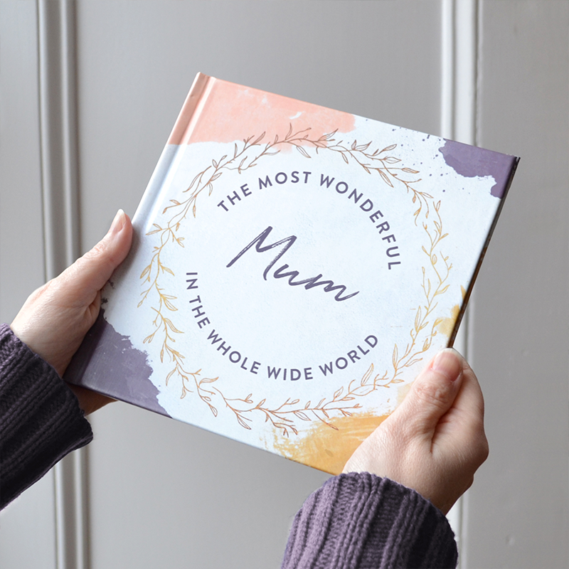 Lady holding a book with pink, purple and gold accents titled "The most wonderful mum in the whole world". The book is being held in a home setting by a lady wearing a purple jumper.
