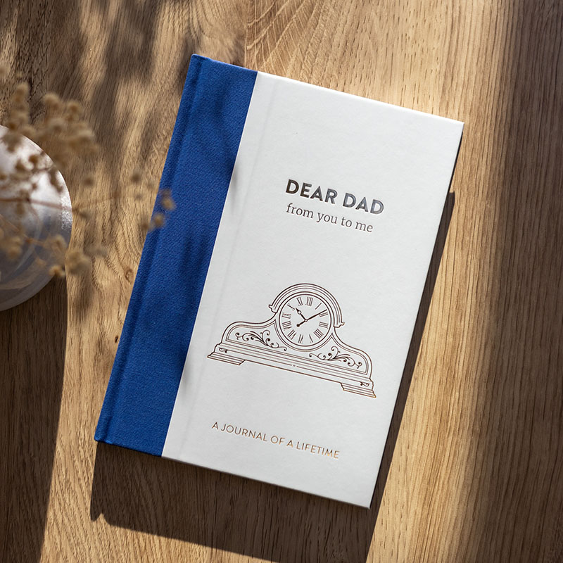 Dear Dad journal on wooden table. The cover features a traditional clock in gold foil, along with the book title. There is a dark blue fabric spine and back. The setting has an autumnal feel.